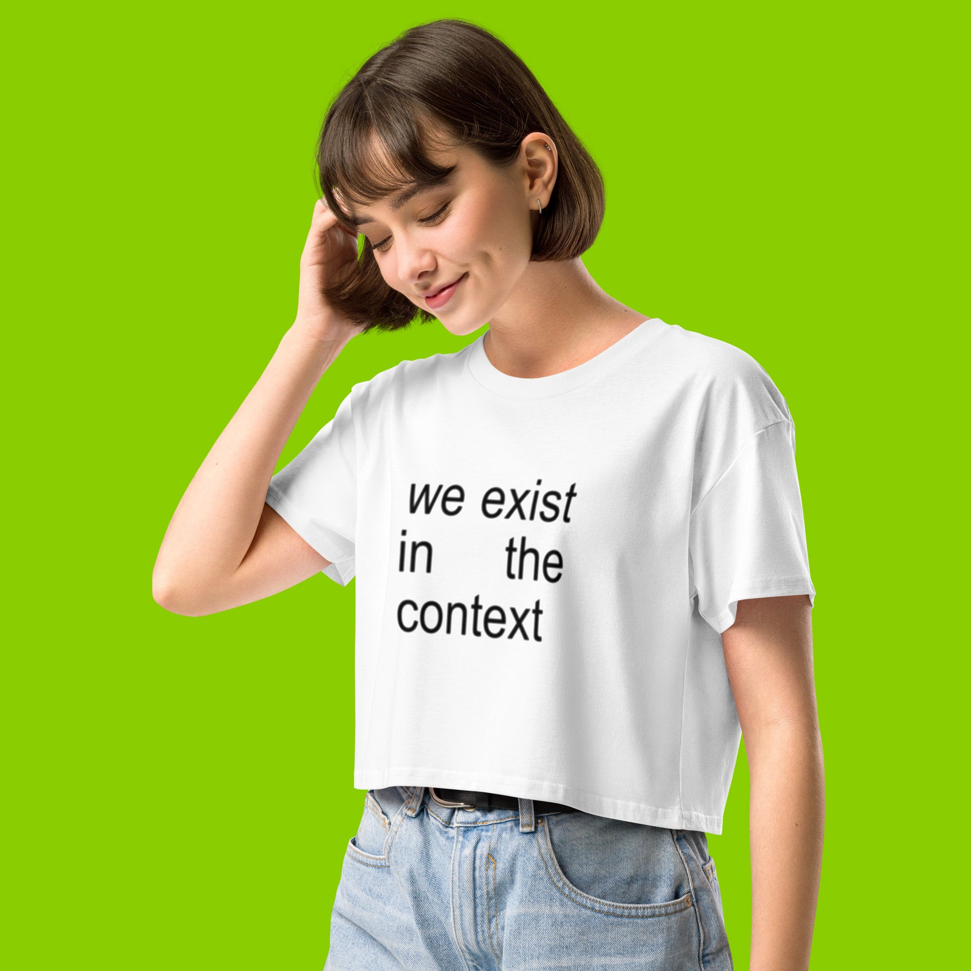 woman wearing white women's crop top t-shirt with the phrase we exist in the context from kamala harris and charli xcx brat