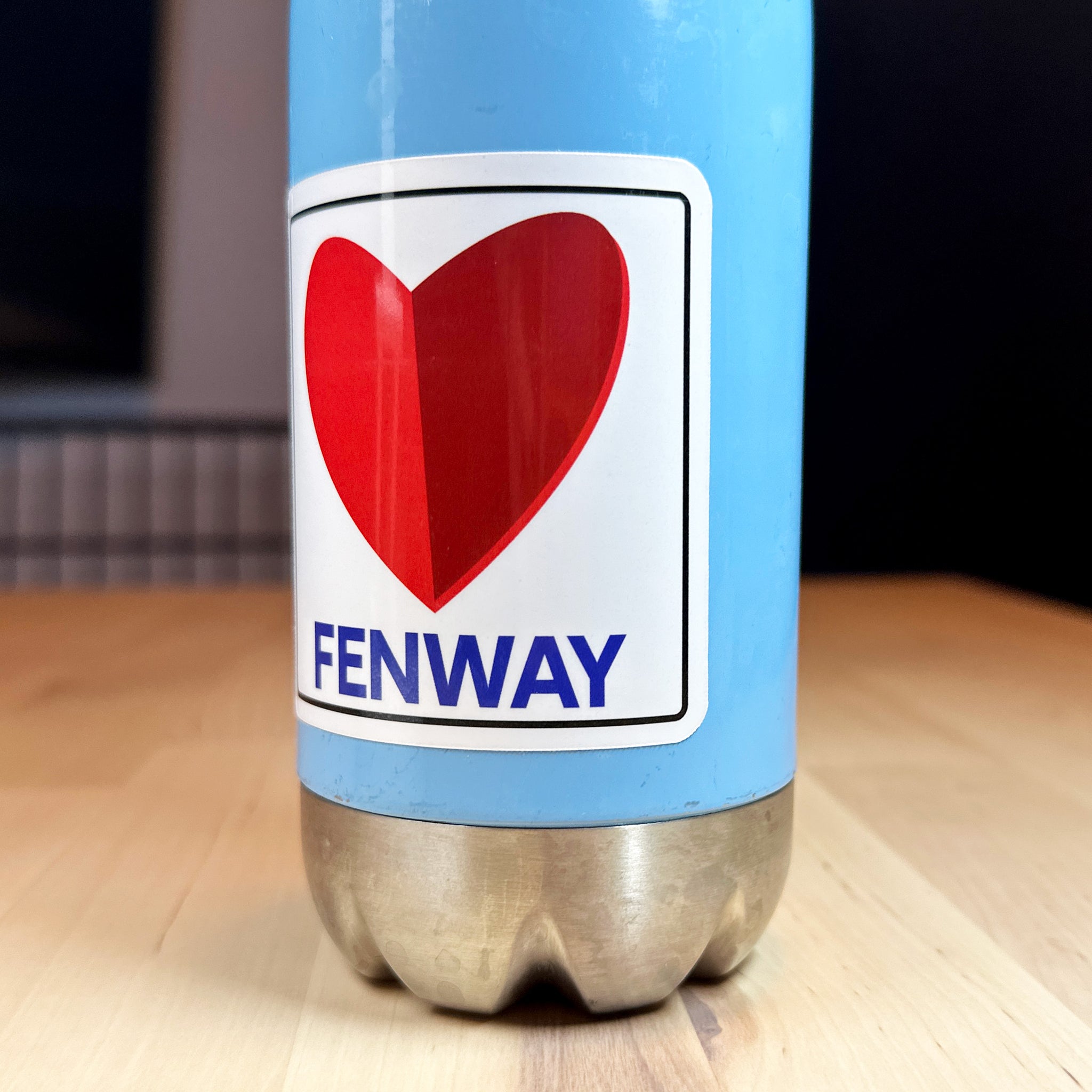 close up photo of a square vinyl sticker with the word FENWAY and a heart in the style of the boston citgo sign on a blue metal bottle