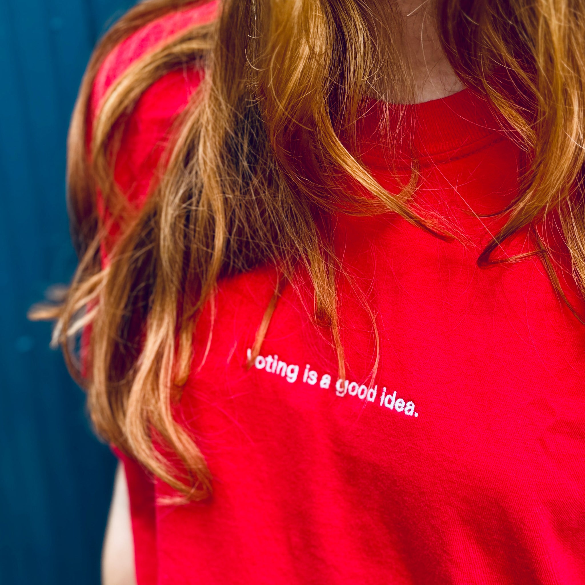 woman wearing red heavyweight unisex t-shirt with the phrase, voting is a good idea, embroidered in white on the front