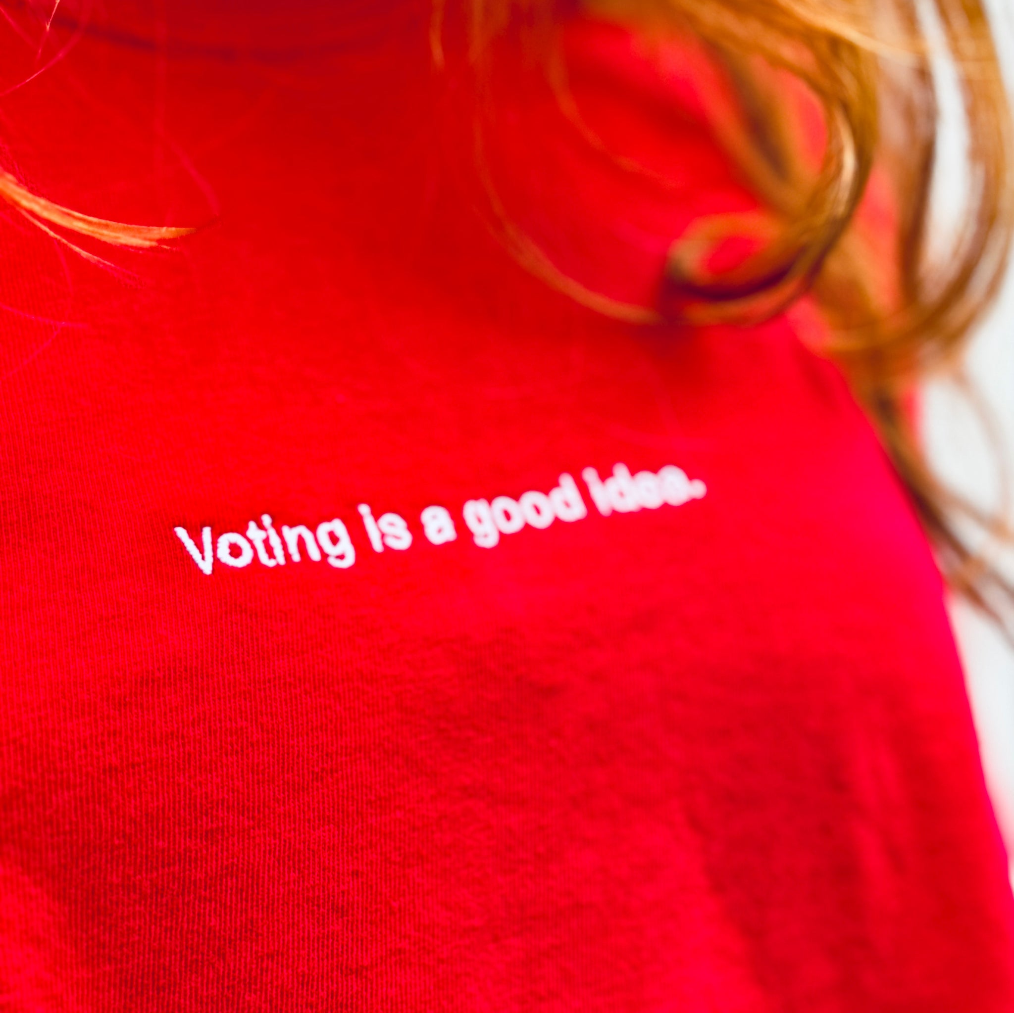 close up of woman wearing red heavyweight unisex t-shirt with the phrase, voting is a good idea, embroidered in white on the front