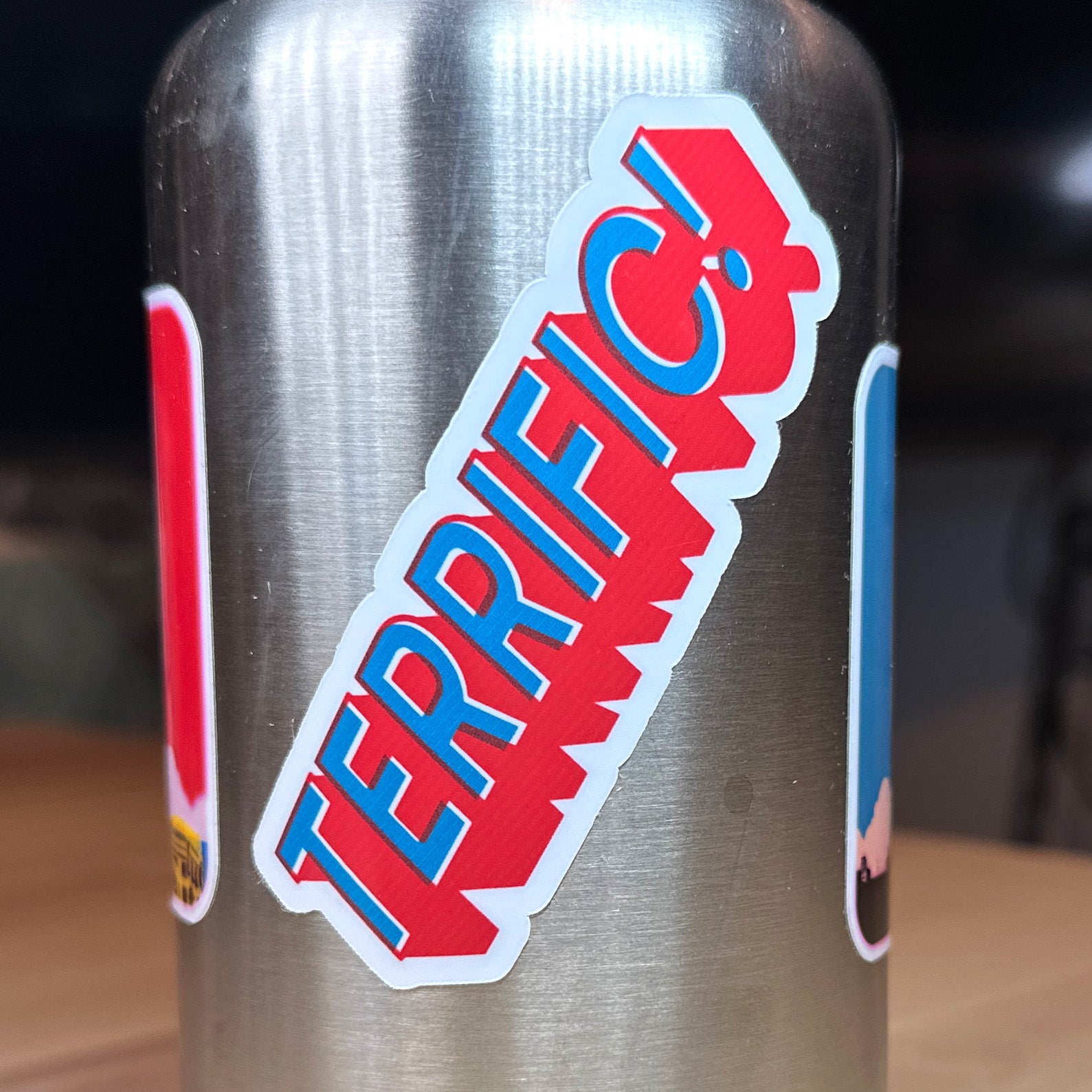 photograph of waterproof sticker of the word Terrific! in teal with a block red outline on a metal water bottle