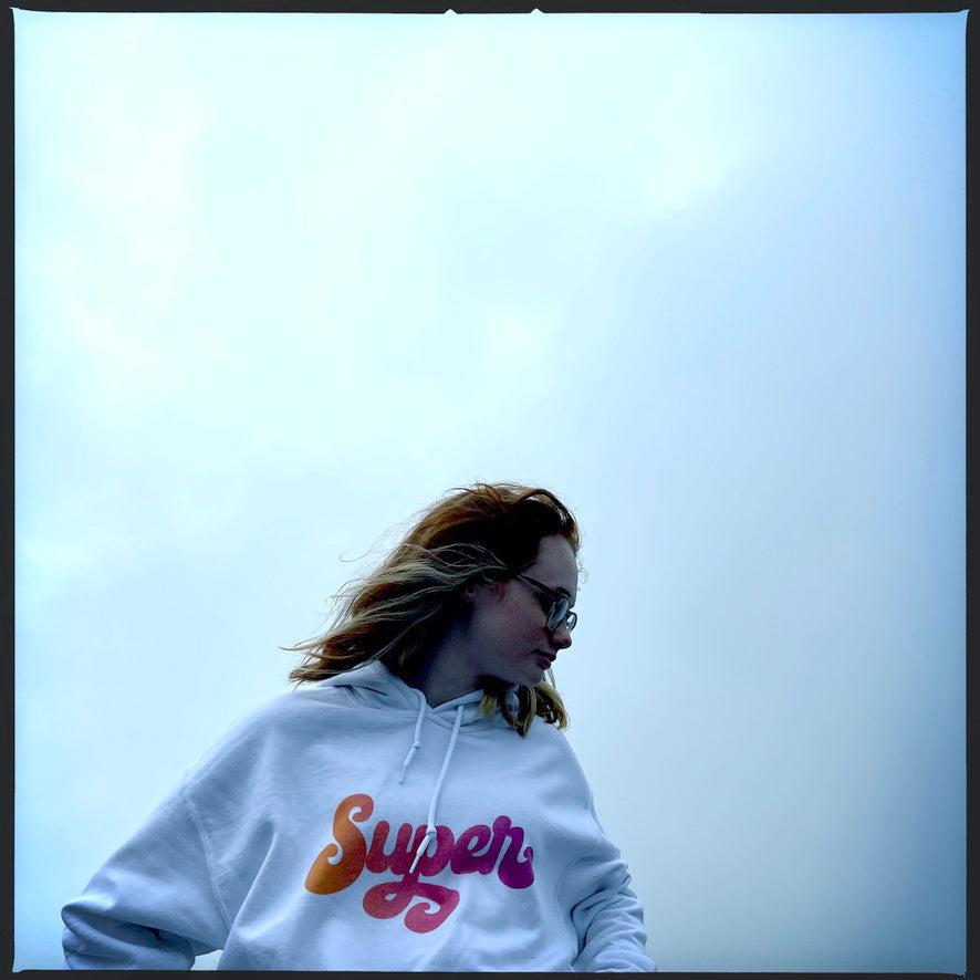 young woman wearing white hoodie with the word Super written in a pink blend cursive lettering